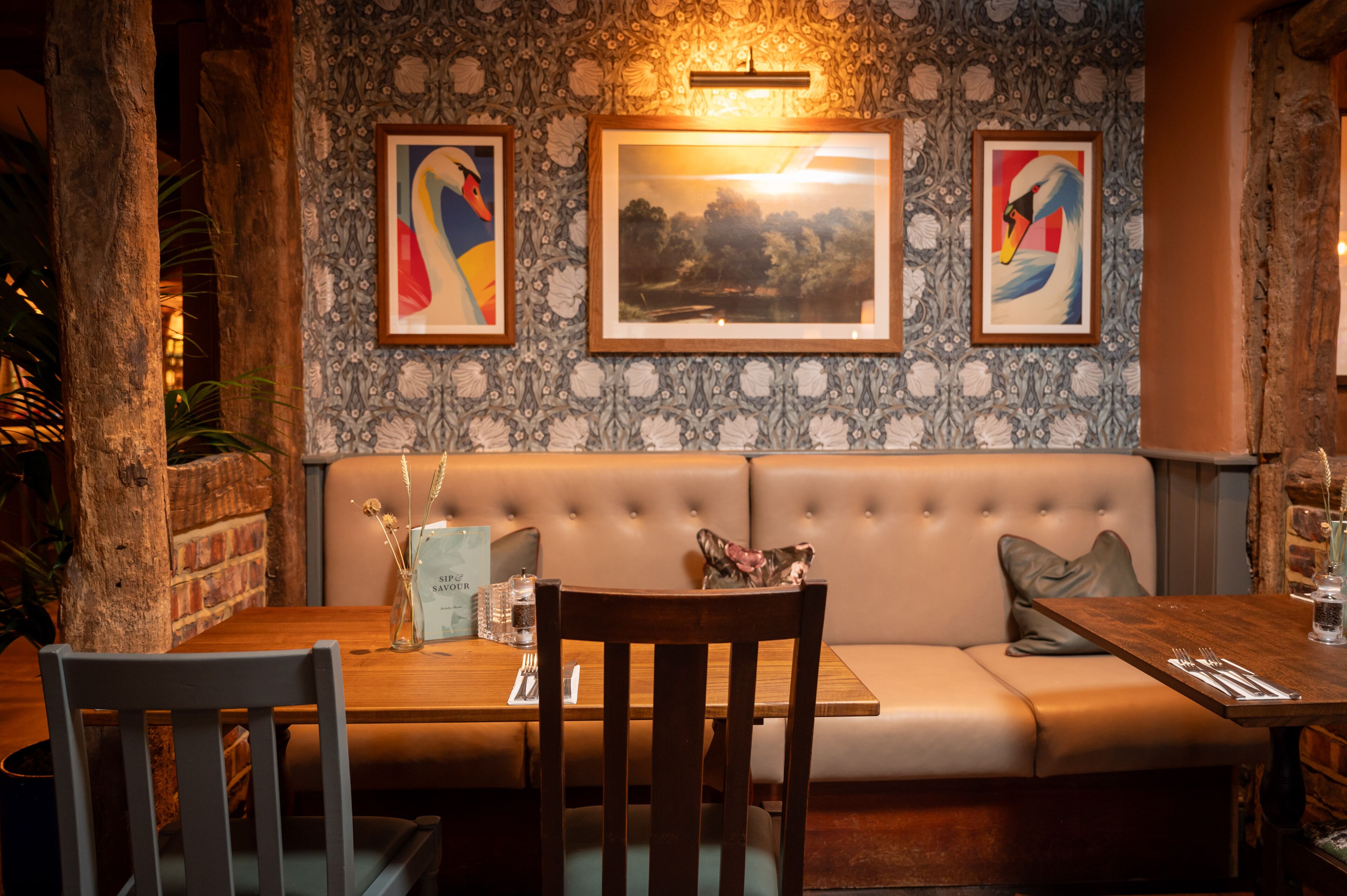 A restaurant interior with a wooden table, cushioned seating, framed artwork, and a brick wall.