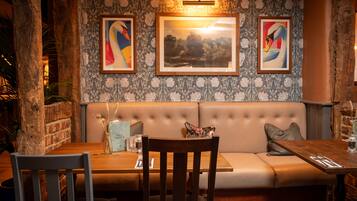 A restaurant interior with a wooden table, cushioned seating, framed artwork, and a brick wall.