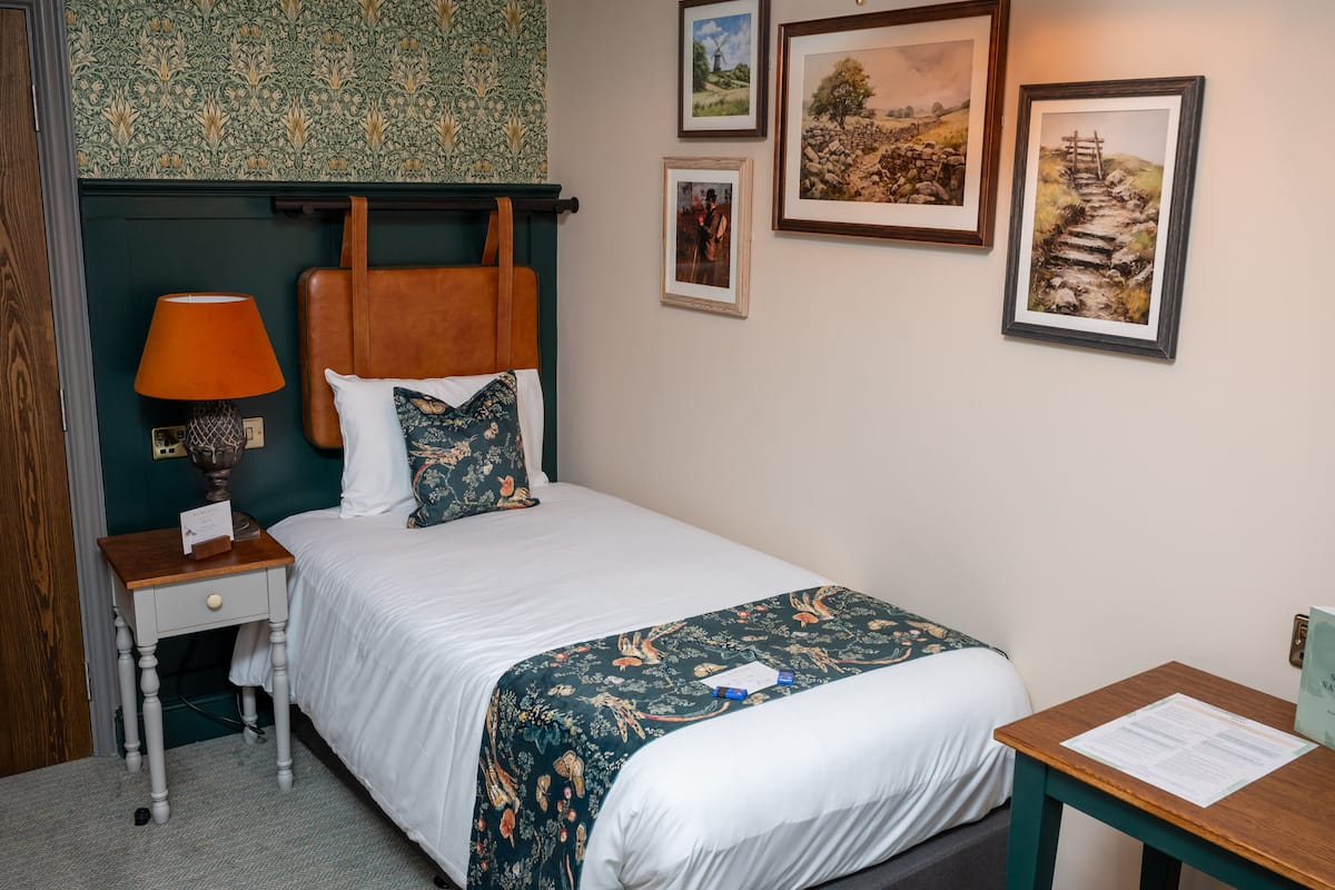 Un letto rifatto con un piumone bianco e una coperta a motivi floreali, un comodino in legno con una lampada, e un tavolino con un menu.