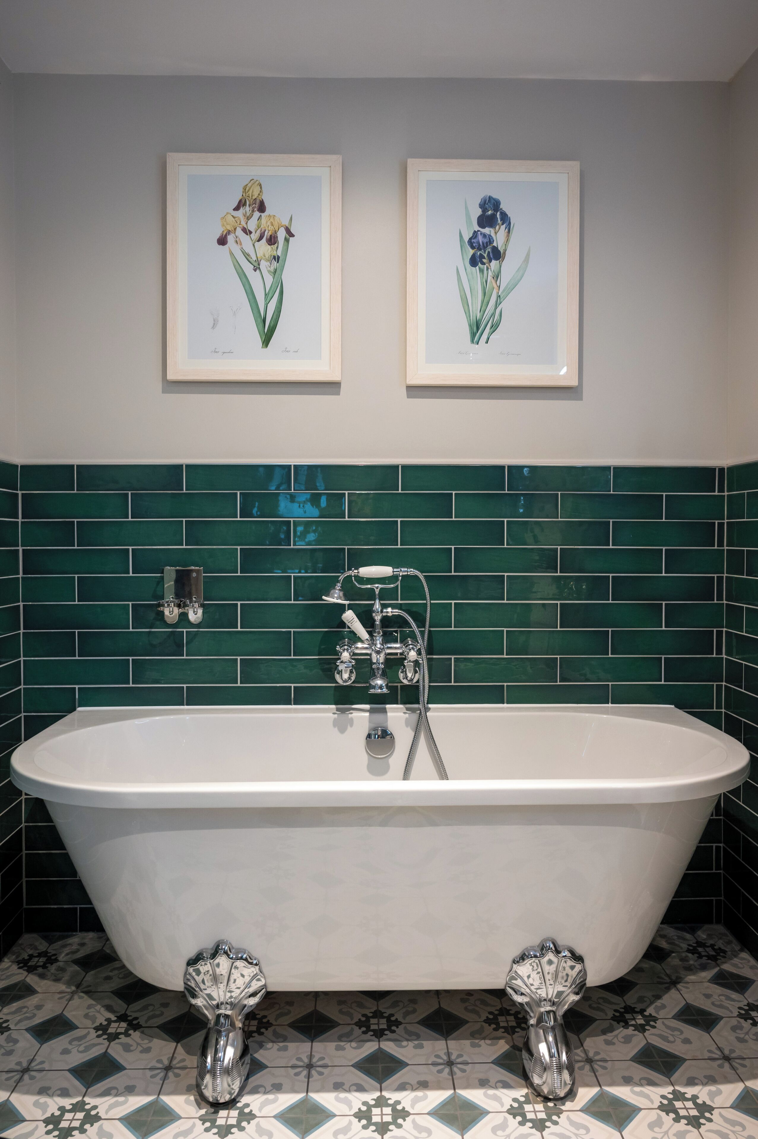 Une salle de bain avec une baignoire sur pieds, des murs recouverts de carreaux verts et deux reproductions botaniques encadrées accrochées au mur.