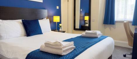 A hotel room with a large bed, a nightstand with a lamp, a mirror, and a window with curtains.