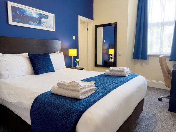 A hotel room with a large bed, a nightstand with a lamp, a mirror, and a window with curtains.