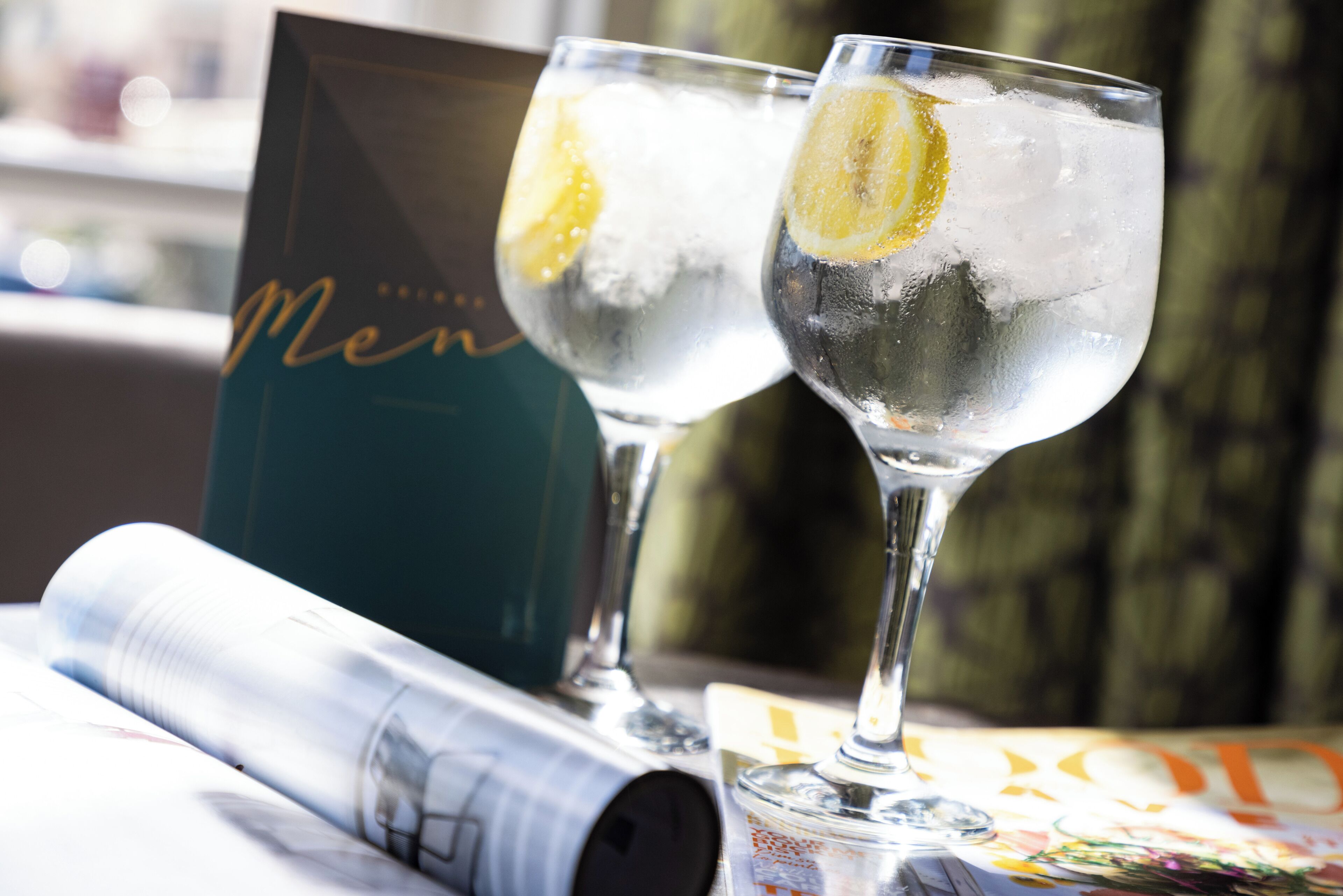 Two glasses of a clear beverage with lemon slices, a book titled 'Men', and a magazine on a table.