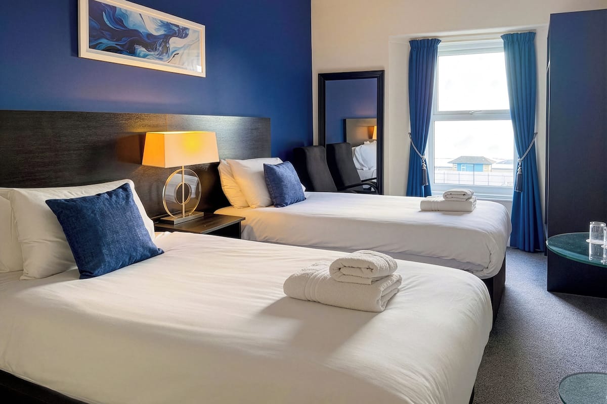 A hotel room with two beds, a nightstand with a lamp, a painting on the wall, and a view of the outside through a window.