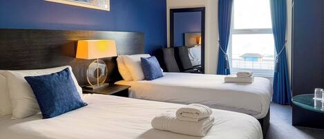 A hotel room with two beds, a nightstand with a lamp, a painting on the wall, and a view of the outside through a window.