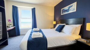 A hotel room with a bed, a nightstand, a lamp, a window with curtains, a painting on the wall, and a small table with a plant. - The Esplanade Hotel by Compass Hospitality (Paignton)