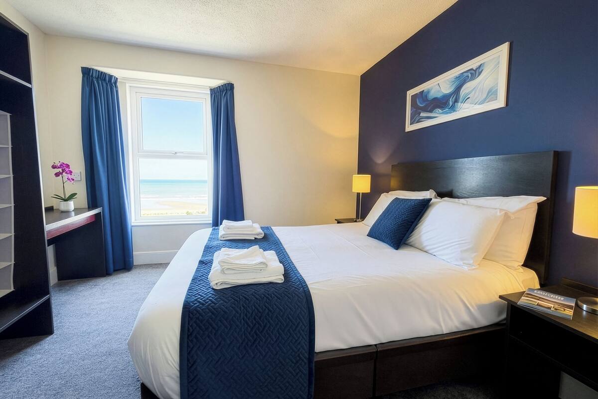 A hotel room with a bed, a nightstand, a lamp, a window with curtains, a painting on the wall, and a small table with a plant.