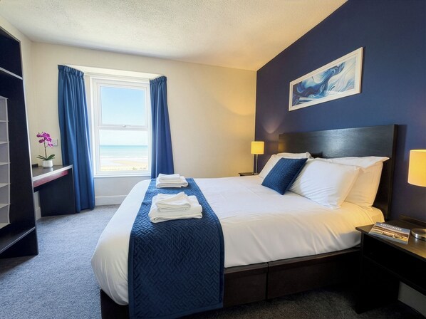 A hotel room with a bed, a nightstand, a lamp, a window with curtains, a painting on the wall, and a small table with a plant.