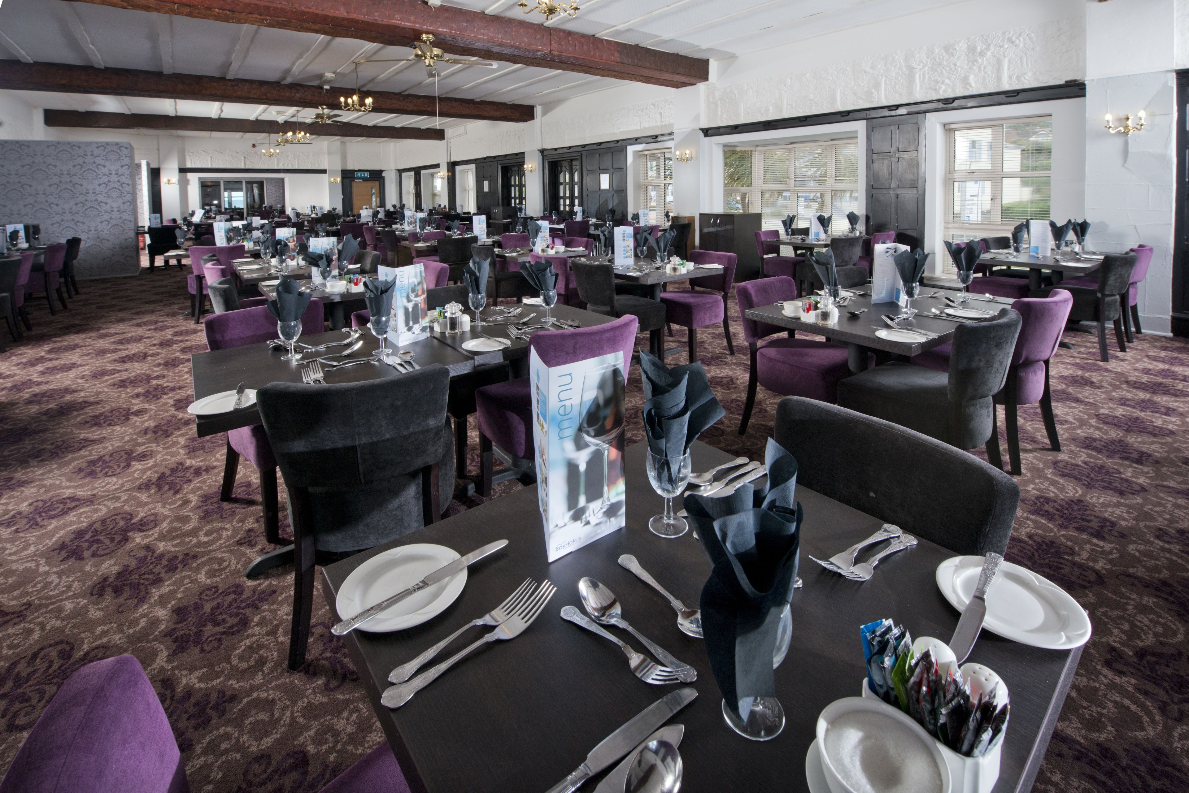 A spacious dining area with multiple tables set for guests, featuring dark tables, purple chairs, and a carpeted floor.