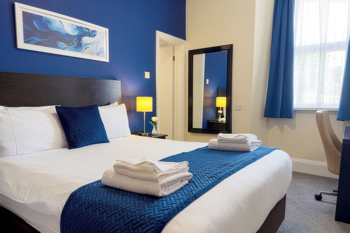 A hotel room with a large bed, a bedside table with a lamp, a mirror, and a window with curtains.