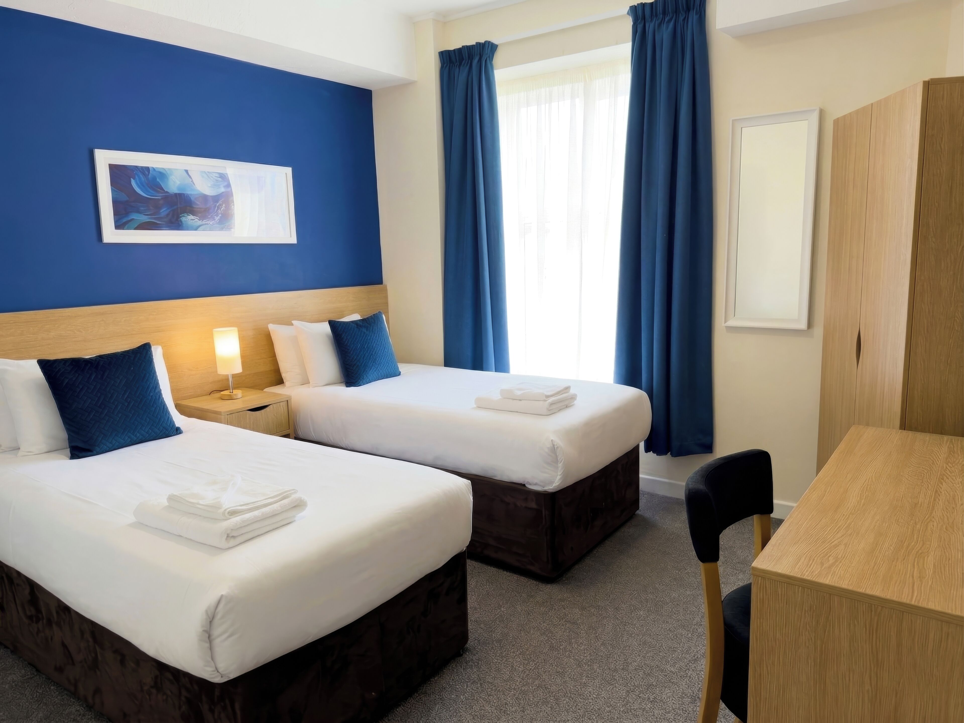 A hotel room with two beds, a desk, a chair, a nightstand, and a painting on the wall.