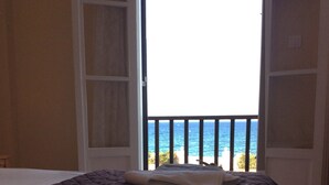 Una cama con una colcha marrón acolchada y dos toallas dobladas. La habitación tiene una puerta abierta que da a un balcón con vistas al océano.