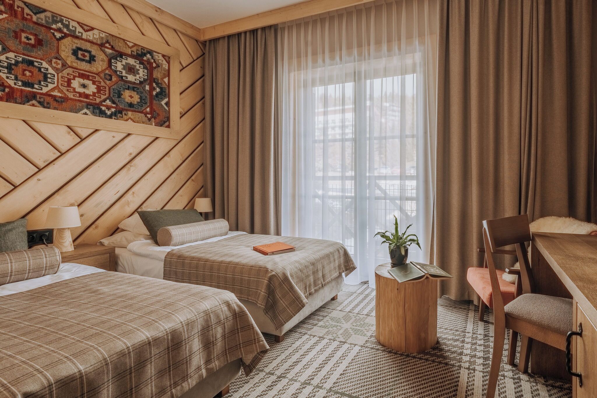 Une chambre d’hôtel avec deux lits, une tête de lit en bois, un bureau, une chaise, une petite table avec une plante et une grande fenêtre avec des rideaux.