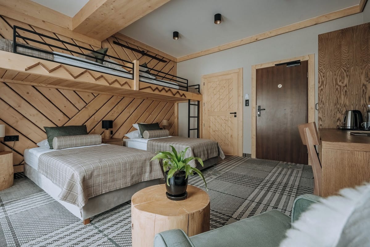A room with two beds, a wooden ceiling, a round wooden table with a plant, and a door with a handle.