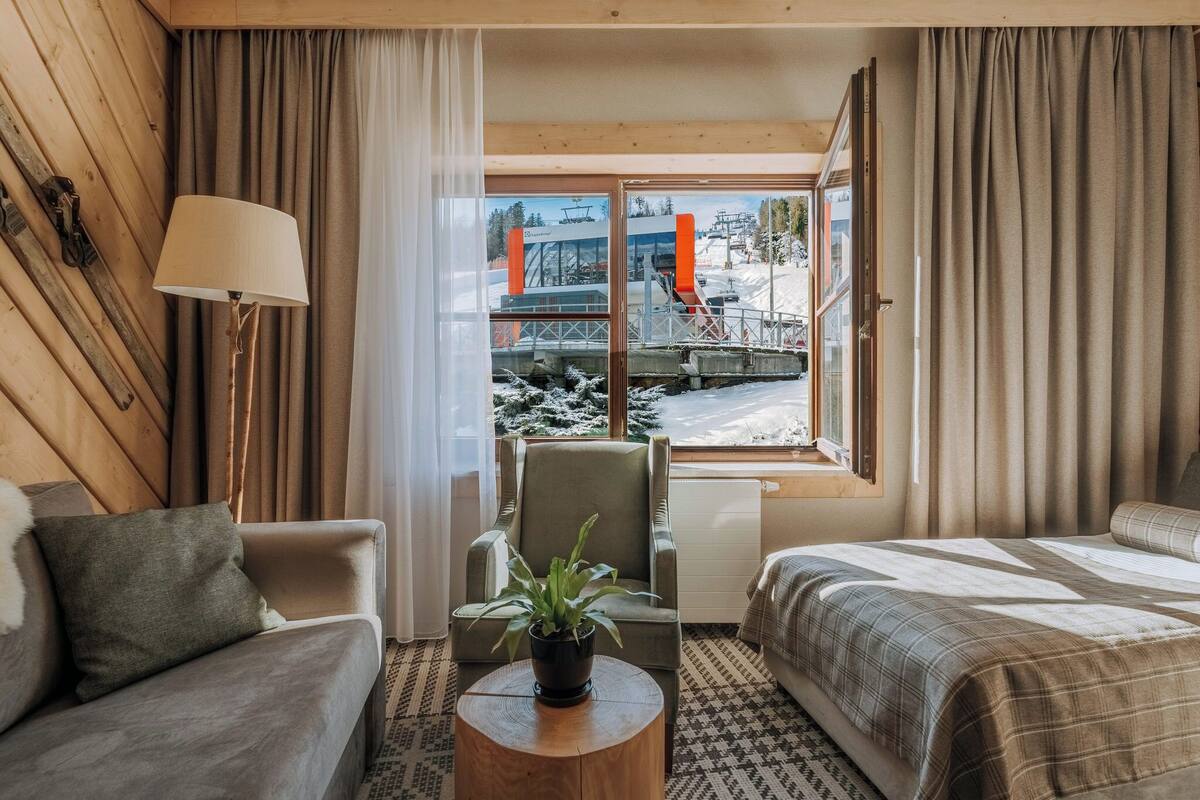 A hotel room with a bed, a sofa, a chair, a lamp, a plant, and a window with a view of a ski lift and snowy landscape.