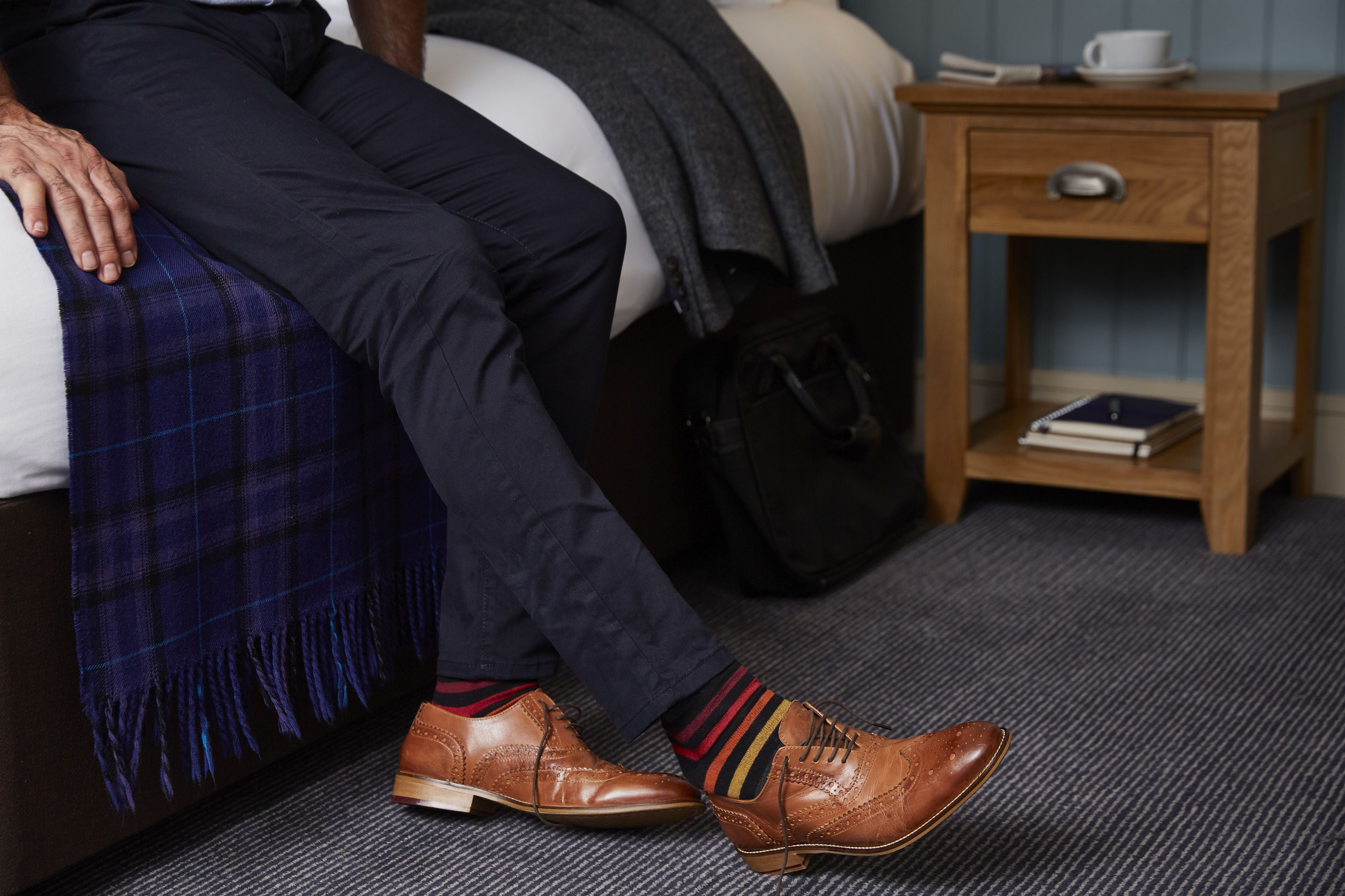 A person sitting on a bed with a plaid blanket, a wooden nightstand with a book and a cup, and a black bag beside the bed.