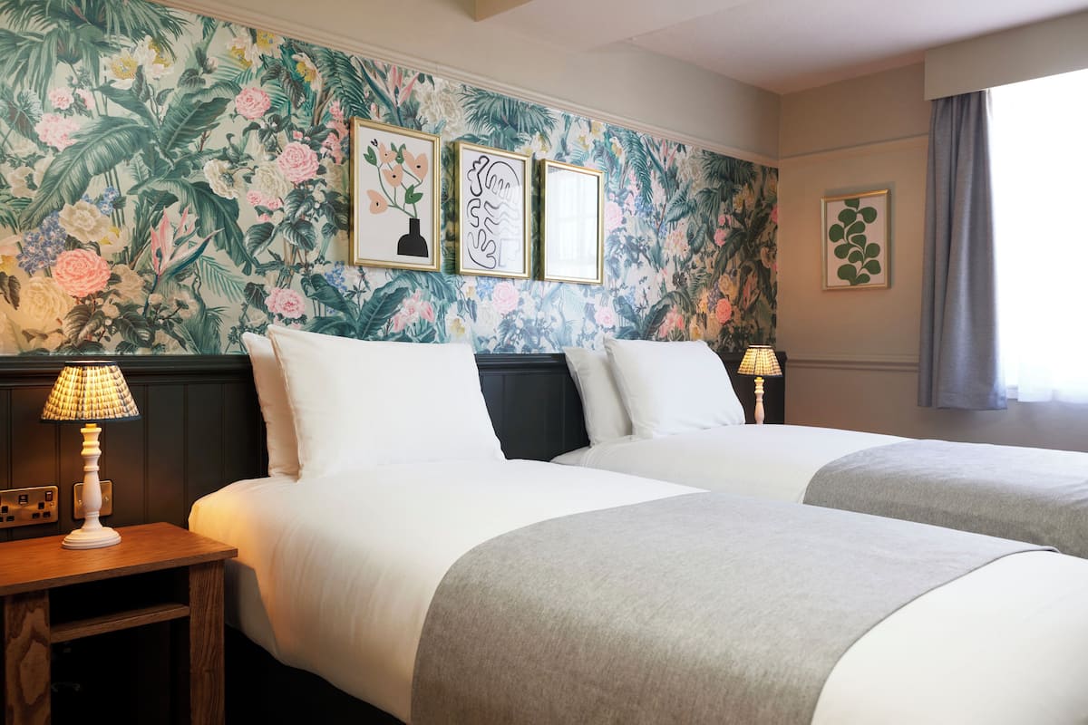 Une chambre d’hôtel avec deux lits, un papier peint à motifs floraux, des œuvres d’art encadrées et des lampes de chevet.