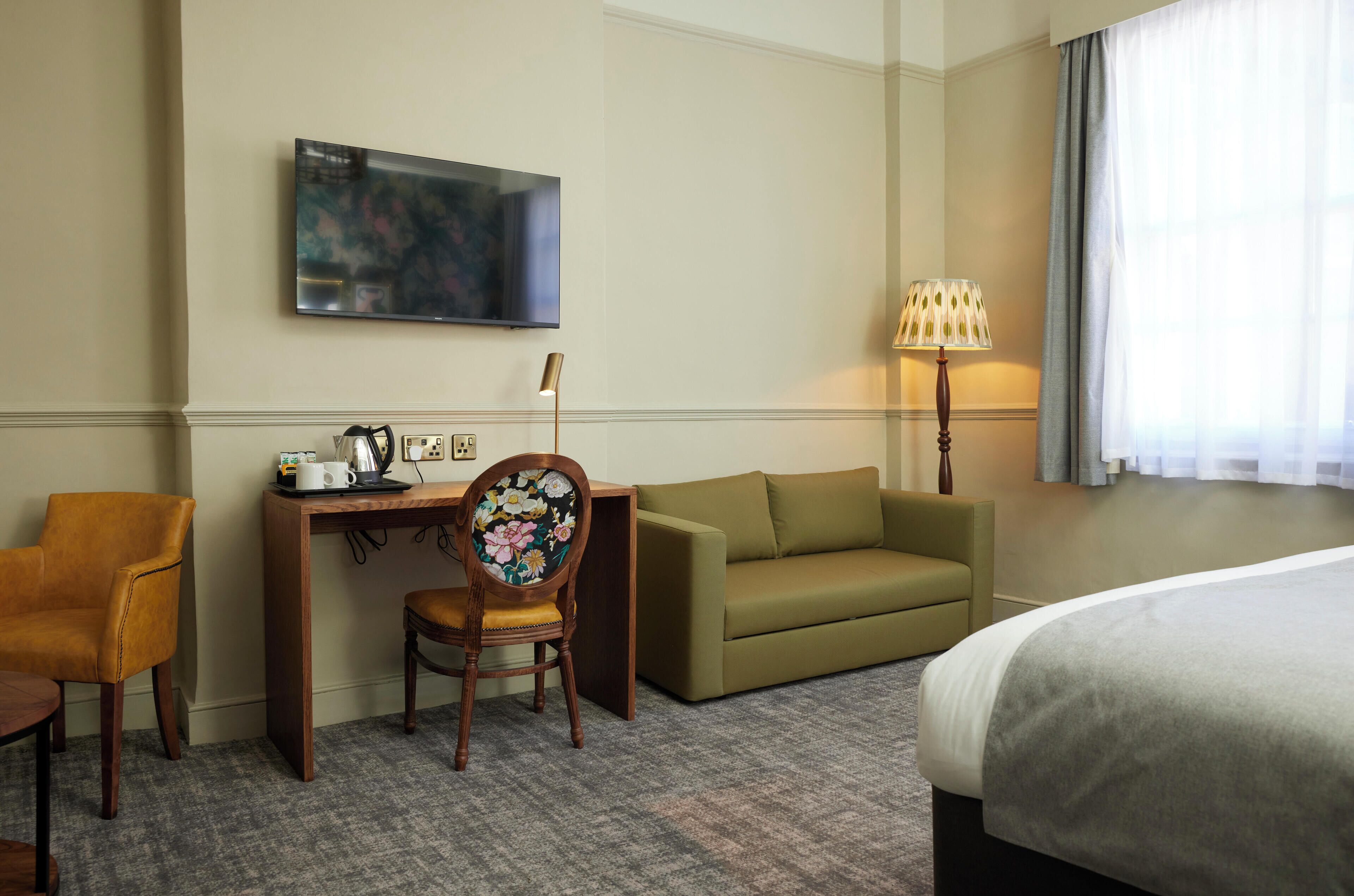 A hotel room with a bed, a desk with a chair, a sofa, a TV mounted on the wall, and a lamp.