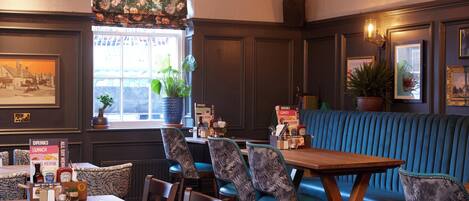 A restaurant interior with a wooden table, blue upholstered chairs, a blue booth seating area, and a window with plants.