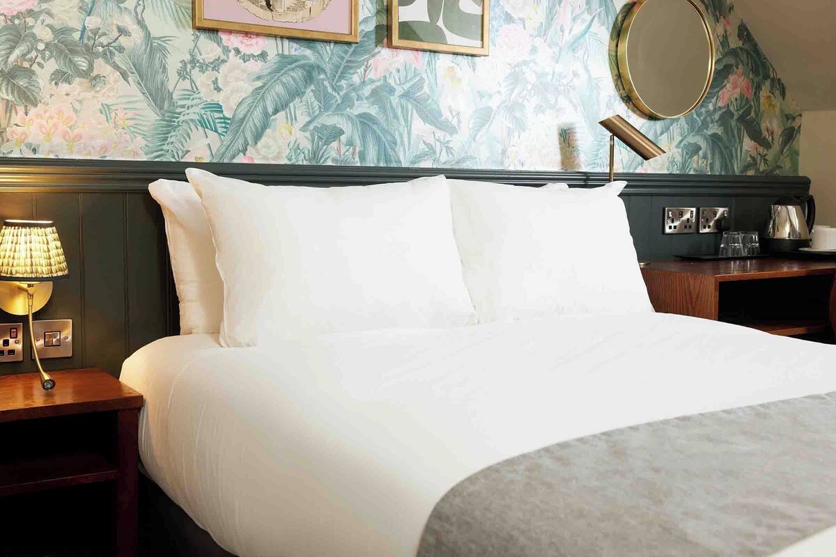 Un lit bien fait, avec du linge de lit blanc, dans une chambre d’hôtel. On y trouve une table de chevet en bois, avec une lampe et une prise de courant. Le mur est décoré d’un papier peint à motifs floraux et d’un miroir rond.