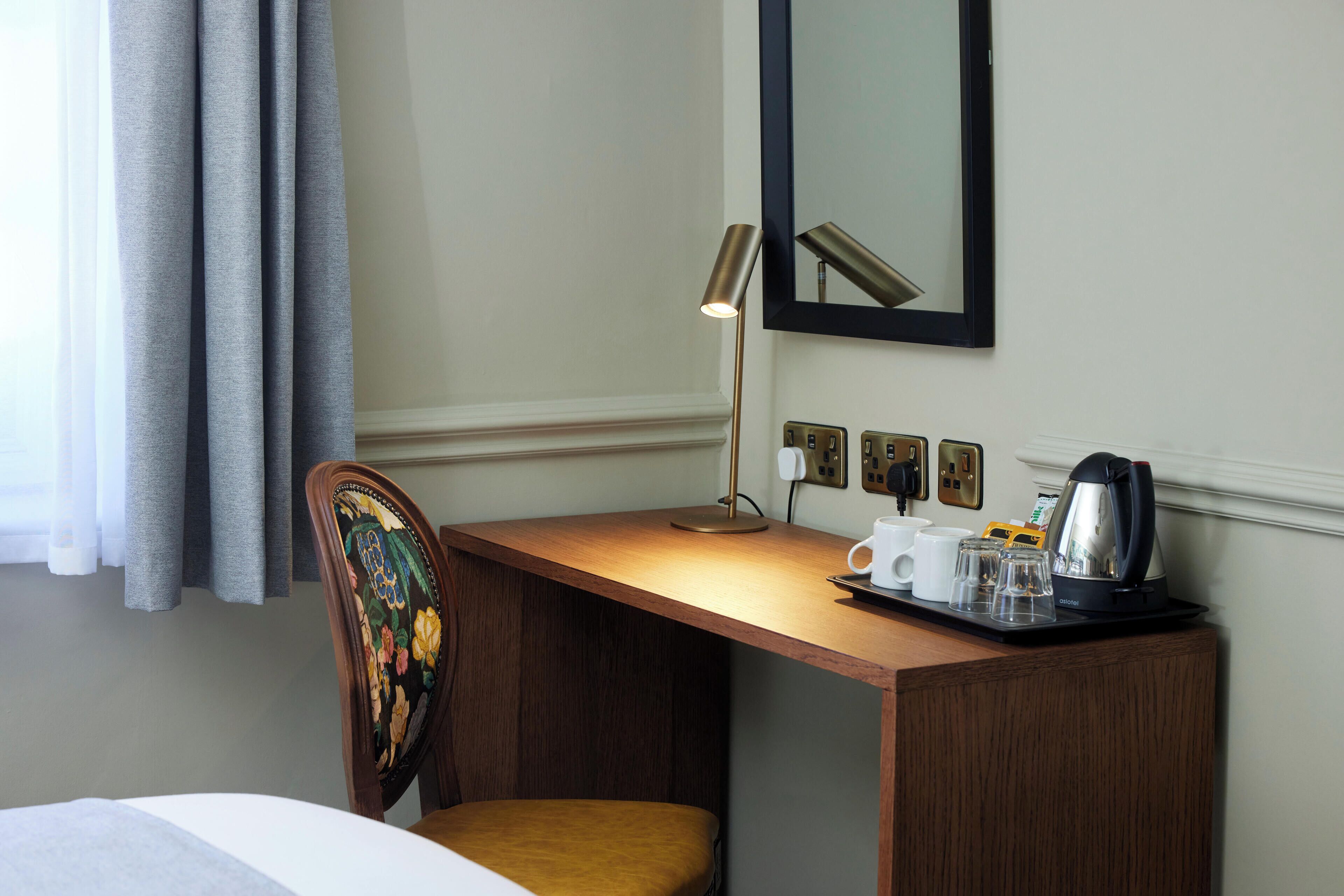 A hotel room with a wooden desk, a chair with floral upholstery, a lamp, a coffee maker, and a tray with a cup and glasses.