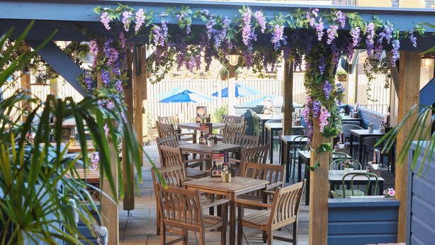 An outdoor patio with wooden tables and chairs, covered by a pergola with purple flowers. - The White Hart Hotel by Greene King Inns (Buckingham)