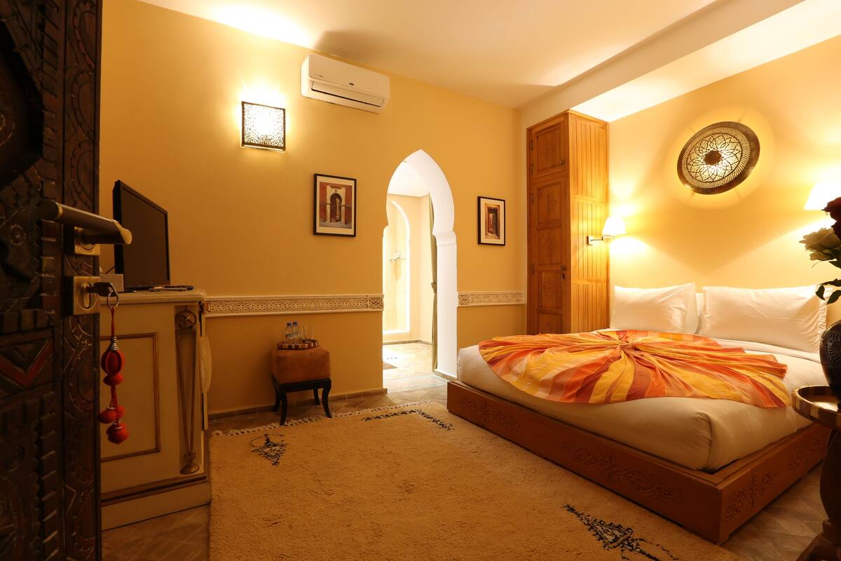 A hotel room with a bed, a desk, an air conditioning unit, and a door with a decorative handle.