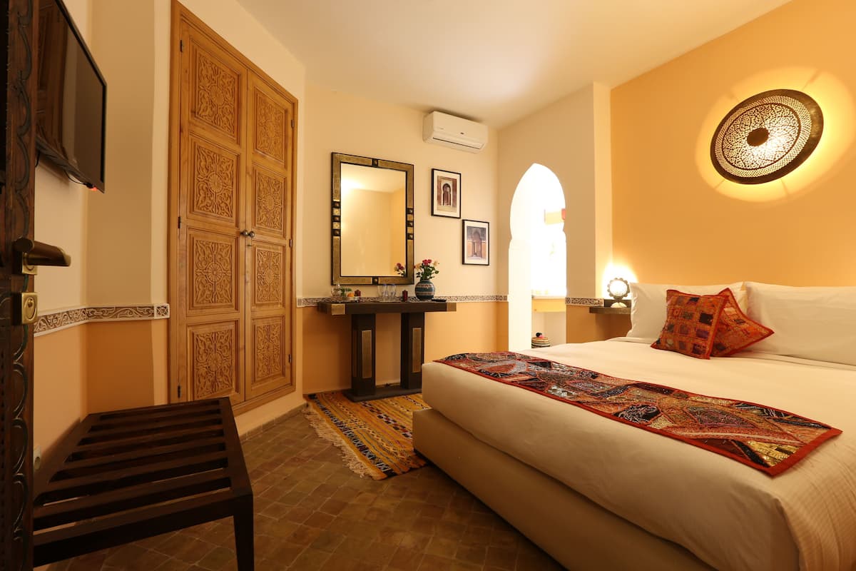 A bedroom with a bed, a wooden wardrobe, a small table, a mirror, and a decorative wall light.