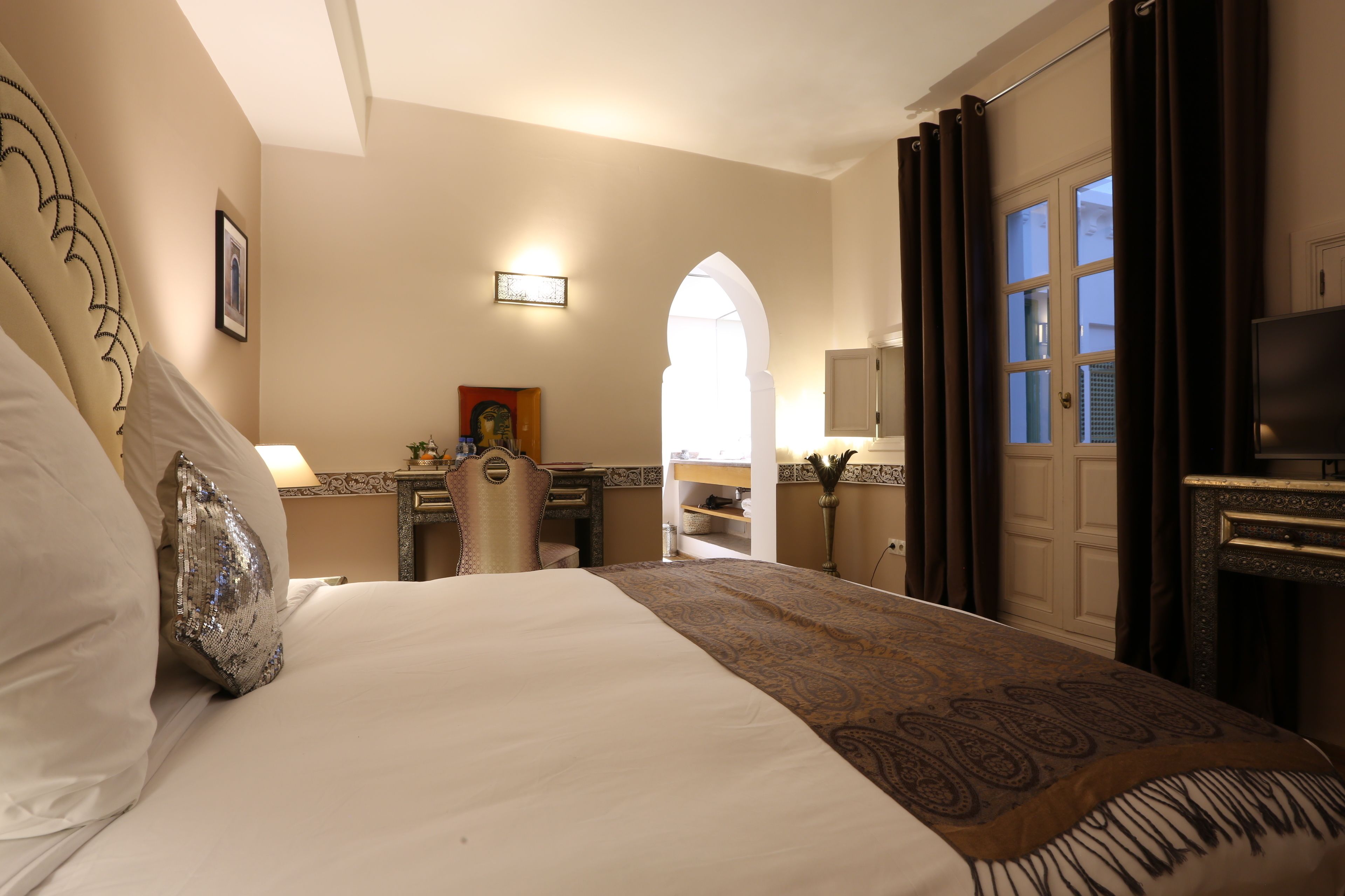 A hotel room with a large bed, a desk, a chair, a television, and a view through an arched doorway.
