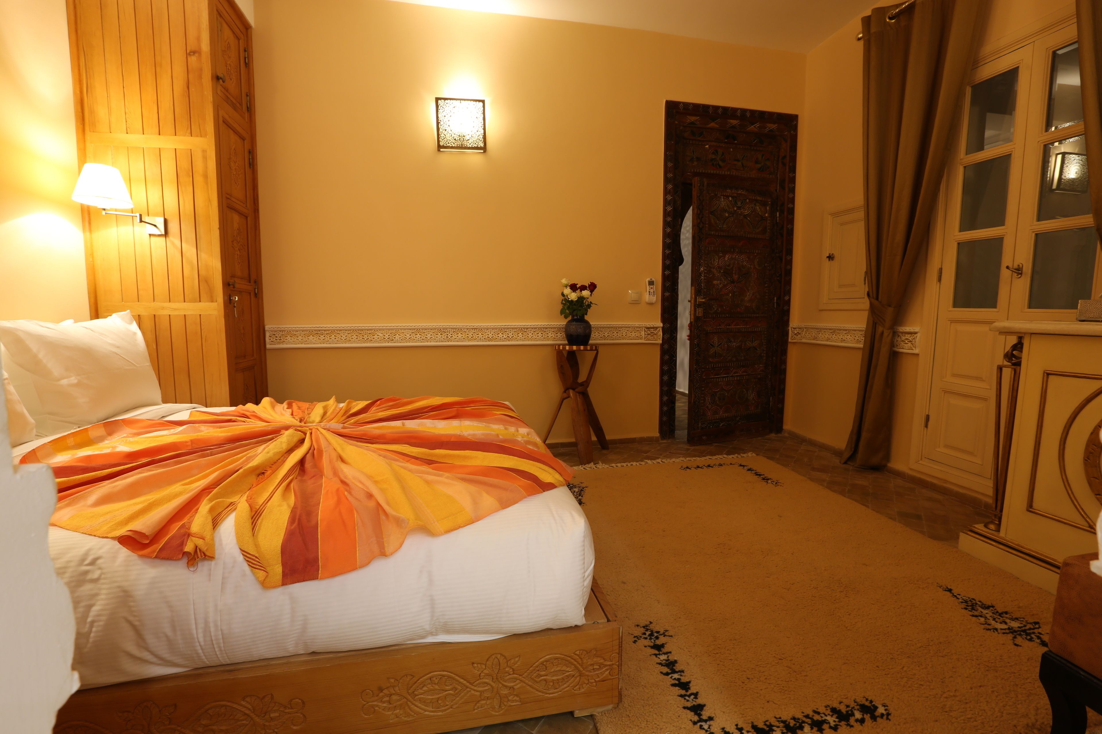 A hotel room with a bed, a nightstand, a lamp, a vase with flowers, and a door.