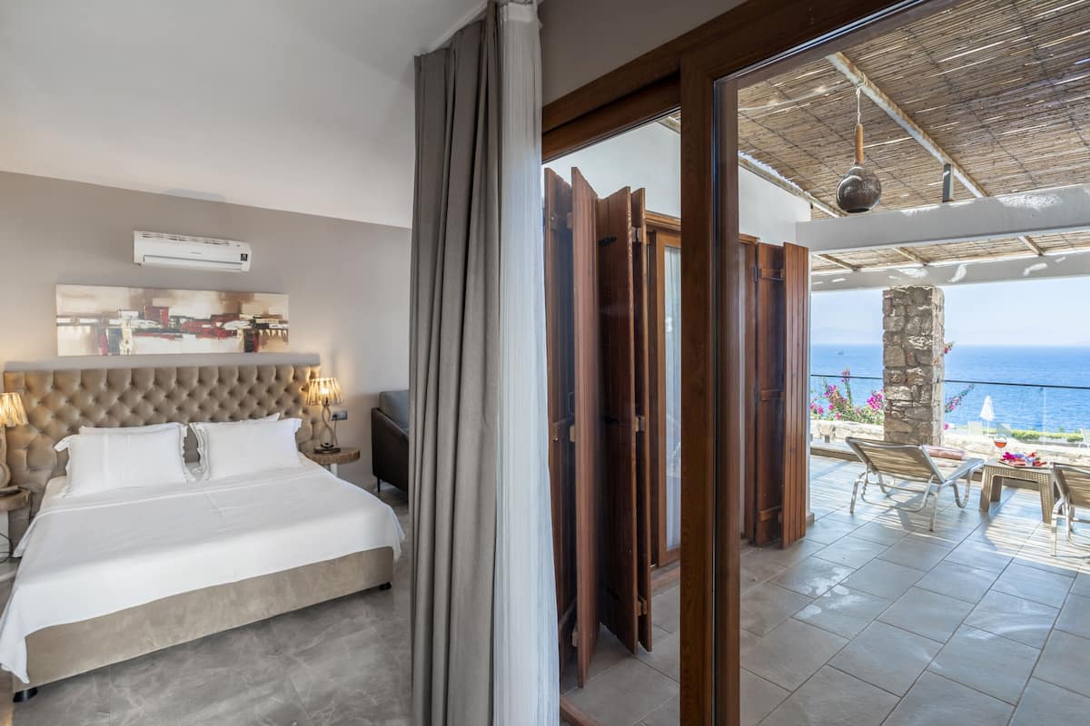 A bedroom with a large bed, bedside lamps, and a view of the ocean through an open door.