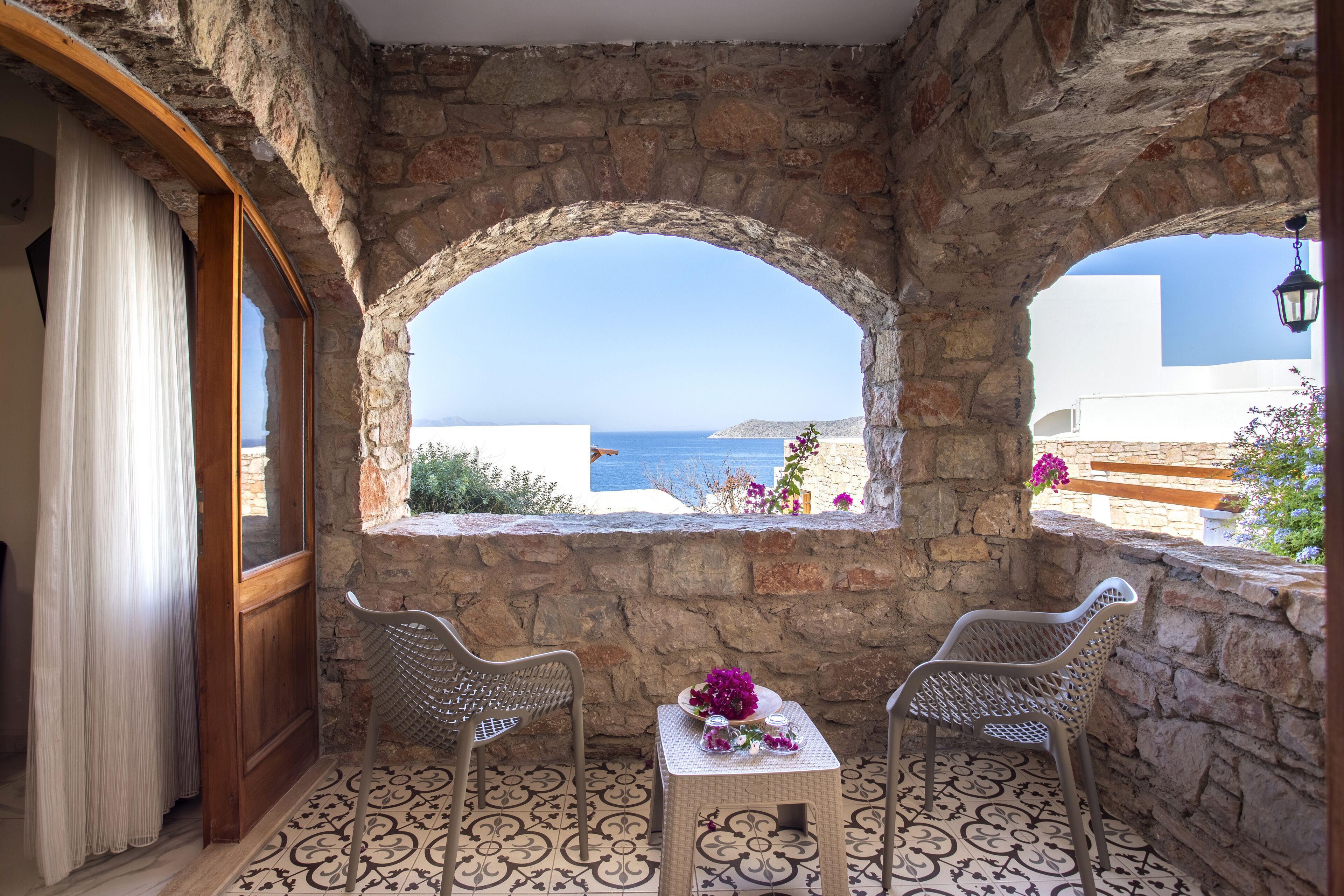 A stone-walled room with two wicker chairs, a small table with a vase of flowers, and a view of the sea through arched windows.