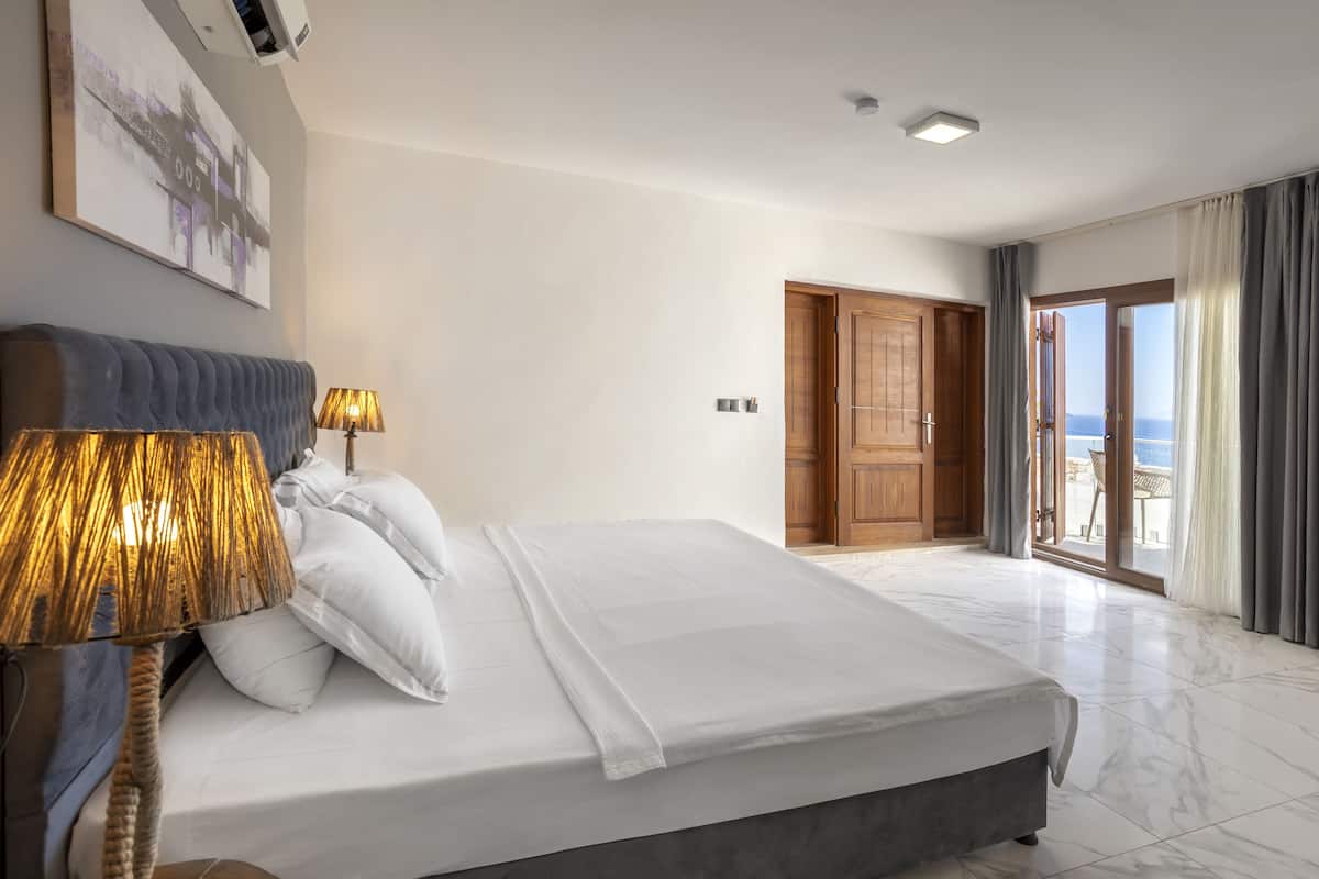 A modern hotel room with a large bed, a bedside lamp, and a view of the sea through a sliding glass door.