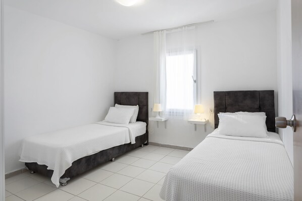 Habitación de hotel moderna y limpia con dos camas individuales, una mesita de noche pequeña con lámpara y una ventana con cortinas traslúcidas.