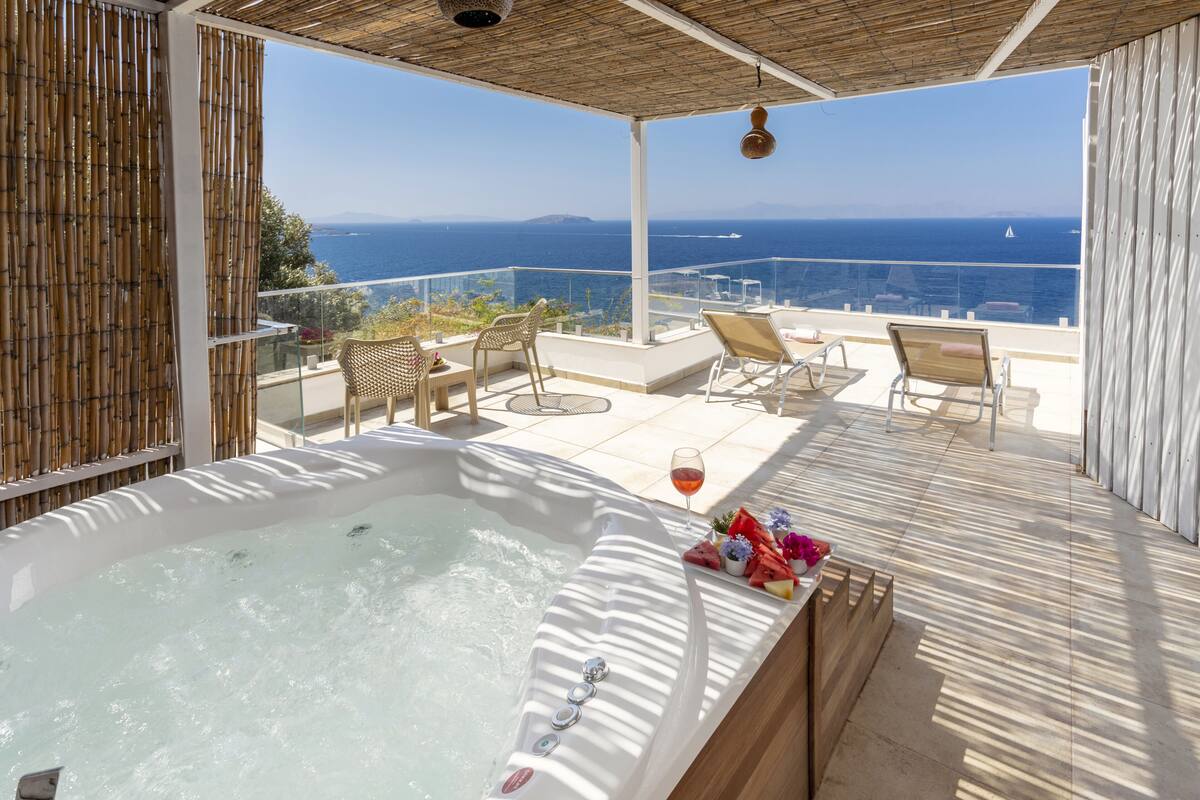 A hot tub on a balcony with a view of the sea, outdoor seating, and a glass railing.