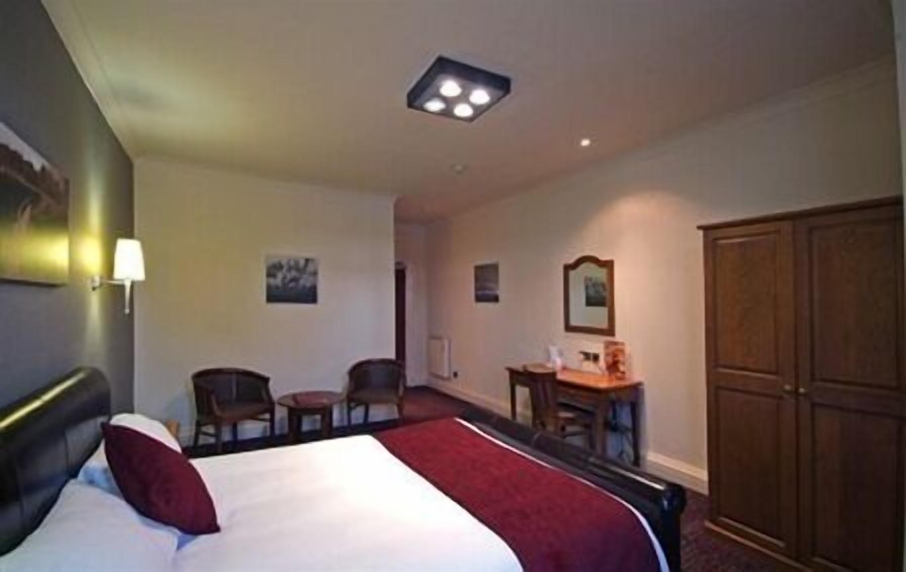 A hotel room with a bed, a desk, two chairs, a wardrobe, and a ceiling light fixture.