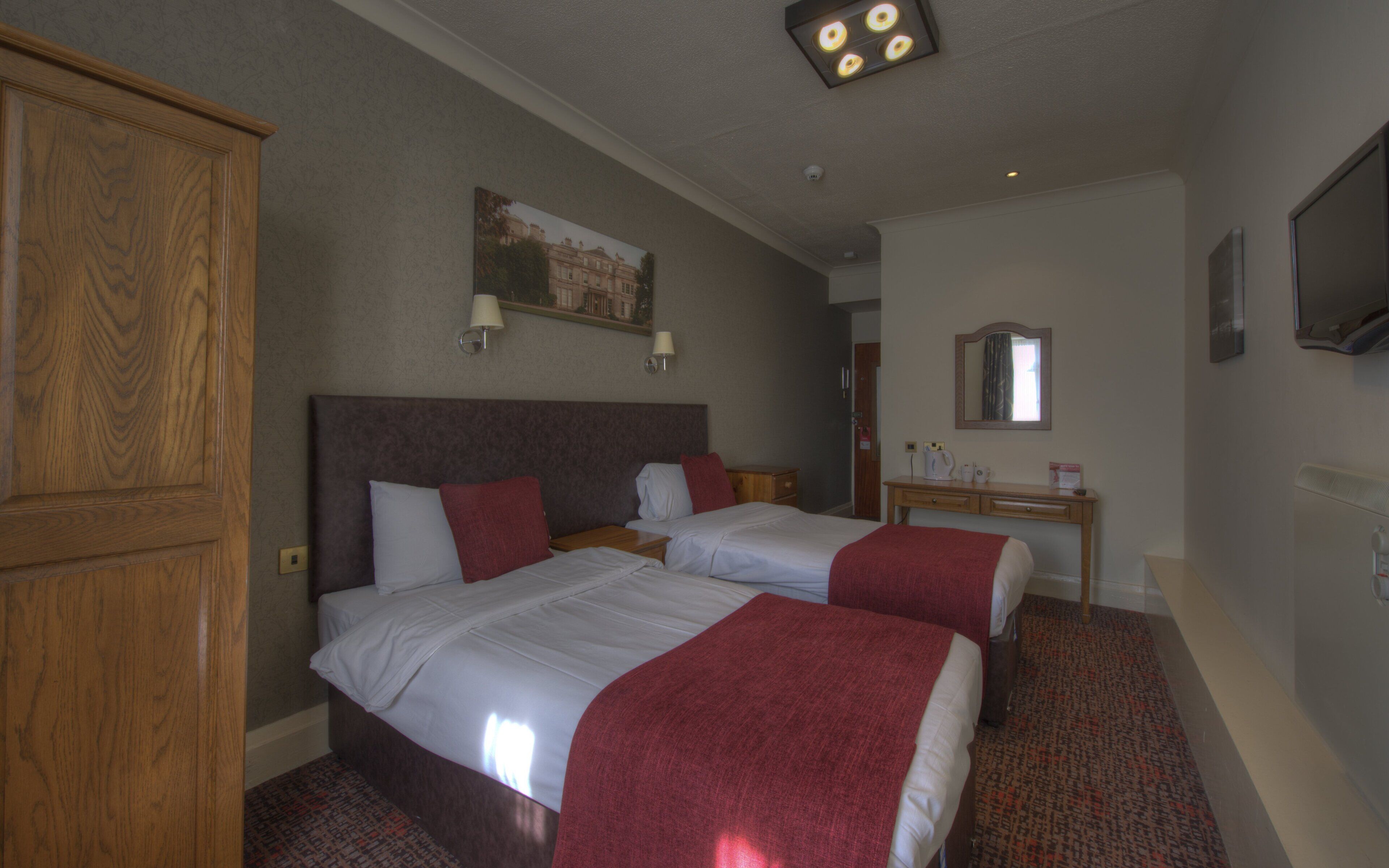 A hotel room with two beds, a wooden wardrobe, a desk with a mirror, and a painting on the wall.
