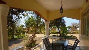 Pátio externo com mesa e cadeiras, rodeado por um jardim e vista para a rua.