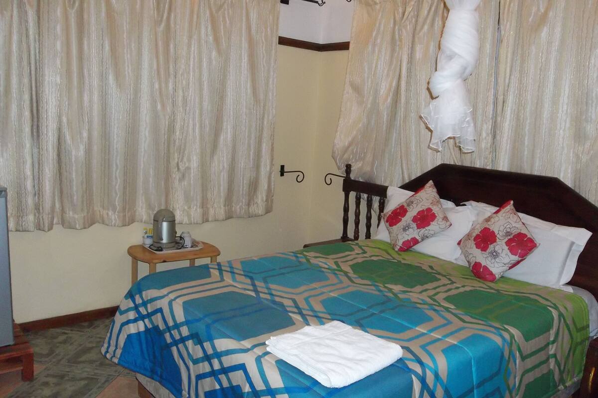A neatly made bed with a patterned bedspread, a bedside table with a kettle, and curtains on the window.