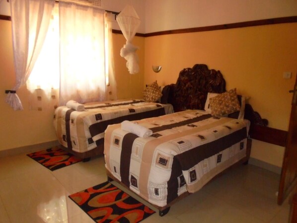 A hotel room with two beds, a large window with curtains, a decorative wall hanging, and a patterned rug.