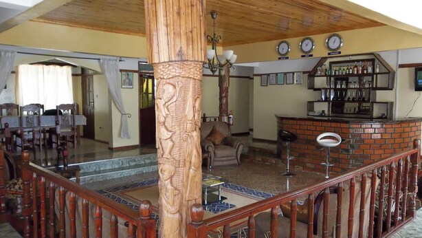 Um hall amplo com teto de madeira, uma coluna central com detalhes entalhados, uma área de bar com prateleiras cheias de garrafas e um espaço de estar com sofá e poltronas.