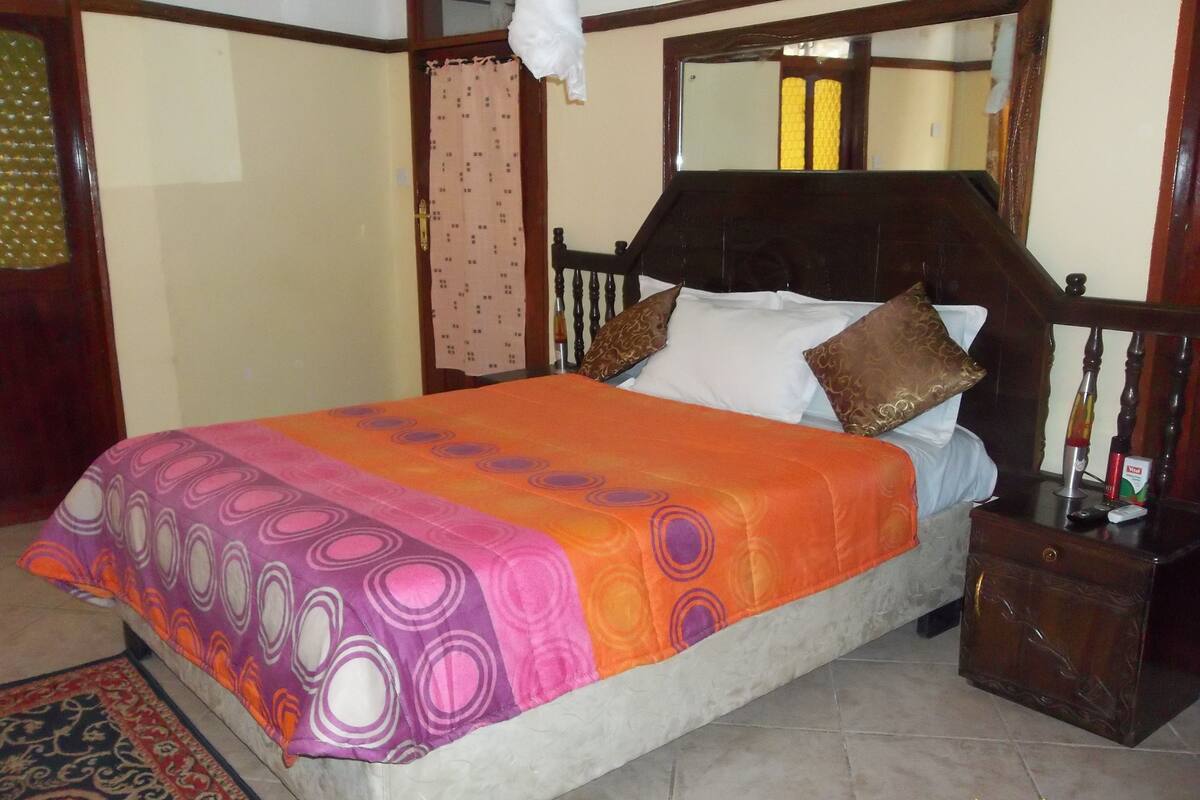 A bed with a patterned bedspread, a wooden headboard, and a nightstand with a bottle and a remote control.
