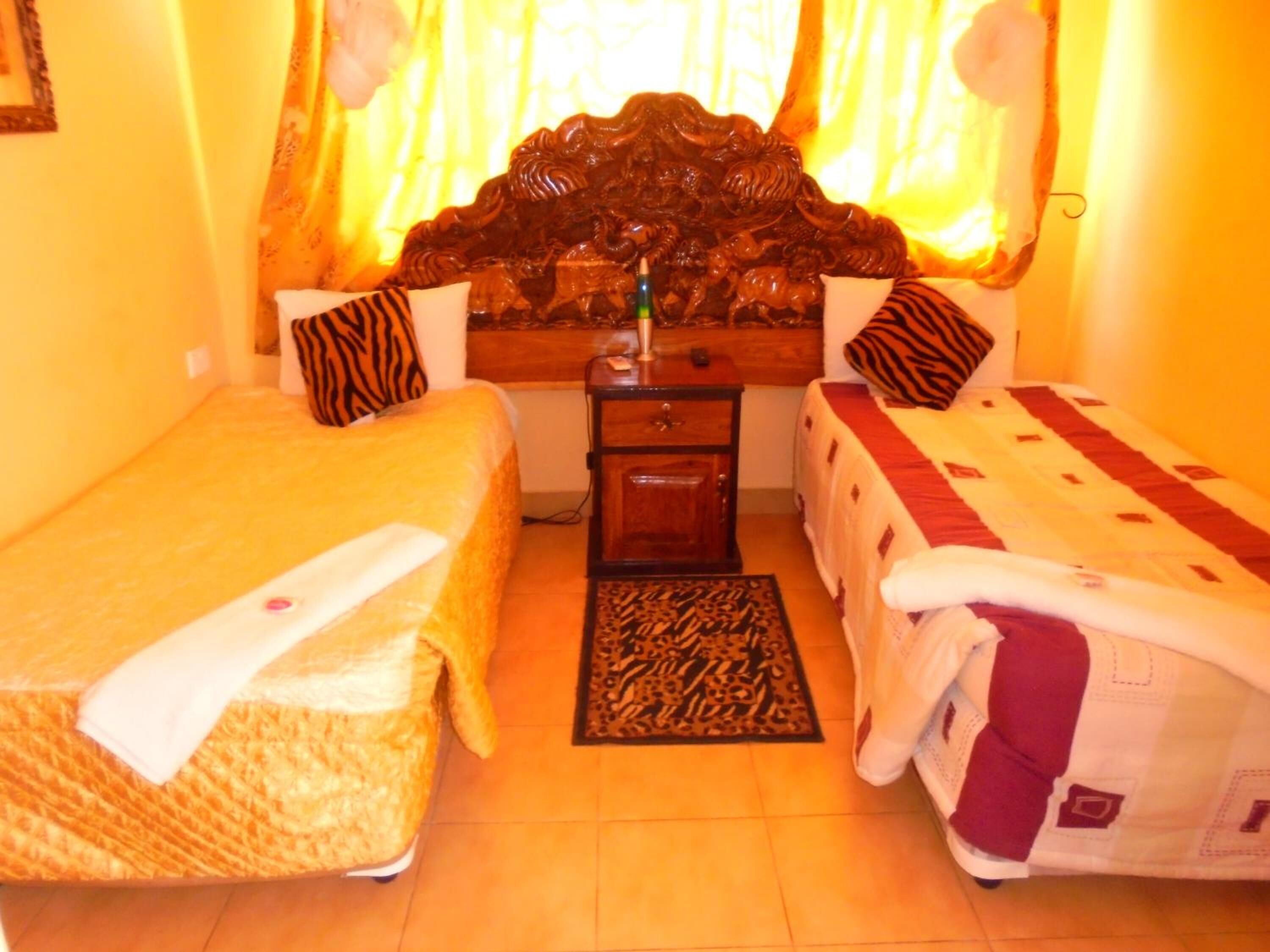 Quarto de hotel com duas camas, cabeceira de madeira, uma mesinha e um tapete estampado.