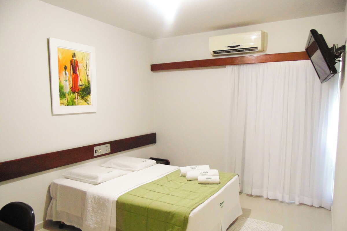A hotel room with a bed, a painting, an air conditioner, and a TV on the wall.