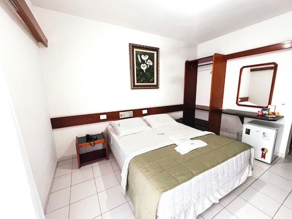 Habitación de hotel con cama, mesita de noche, pequeño escritorio con espejo, refrigerador y un cuadro en la pared.