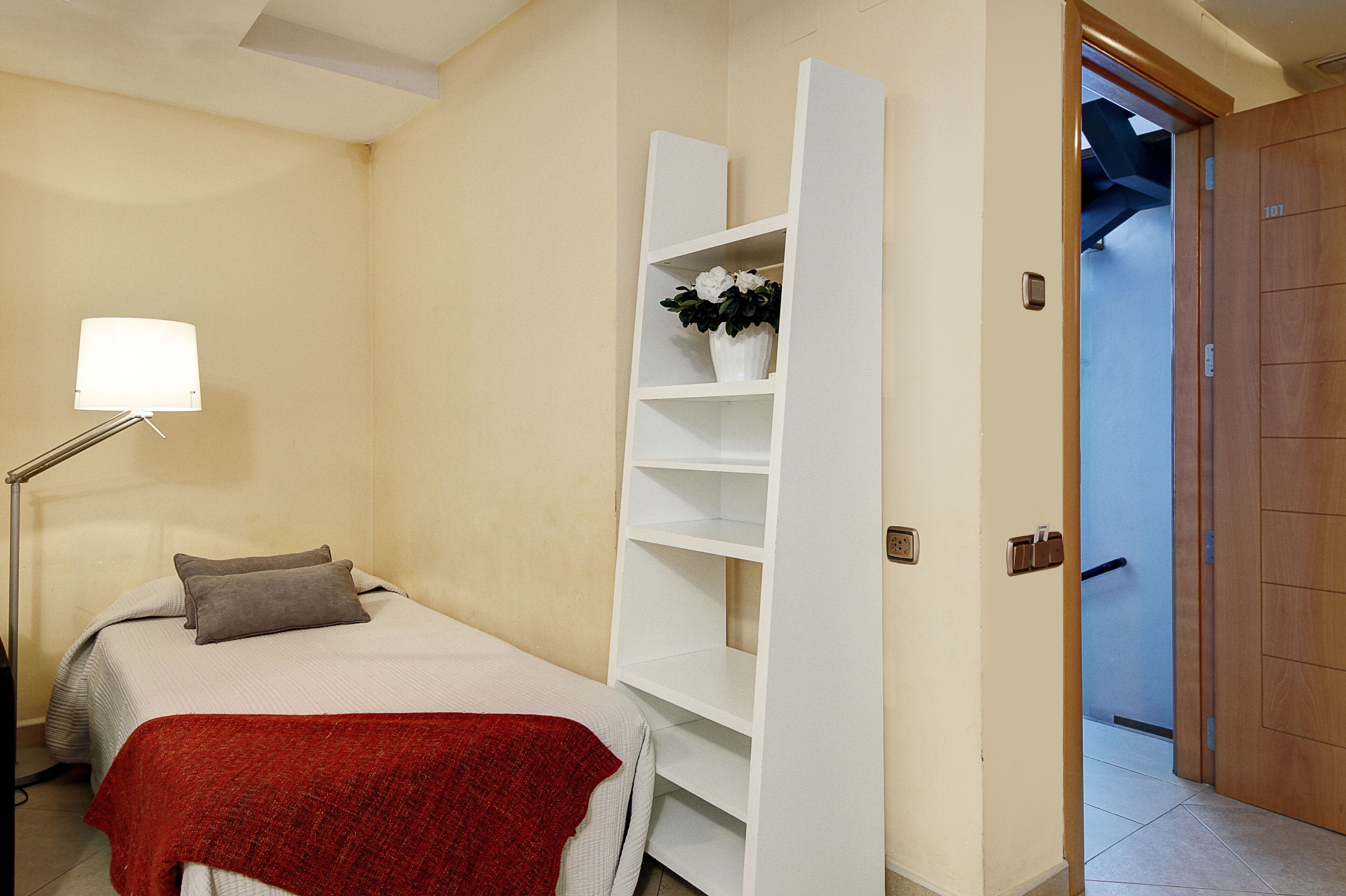 Une chambre à coucher comprenant un lit, une table de chevet, une lampe et une étagère blanche sur laquelle est posée une plante en pot.