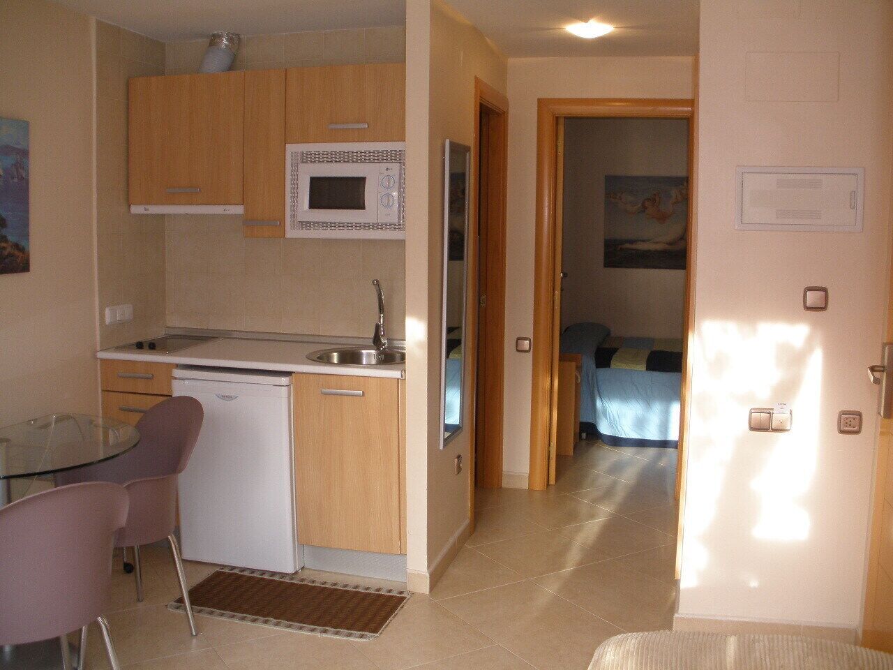 Cocina compacta equipada con microondas, lavamanos y un refrigerador pequeño. Hay una zona de comedor con una mesa de vidrio y sillas. Se ve un dormitorio al fondo, con una cama y un cuadro en la pared.