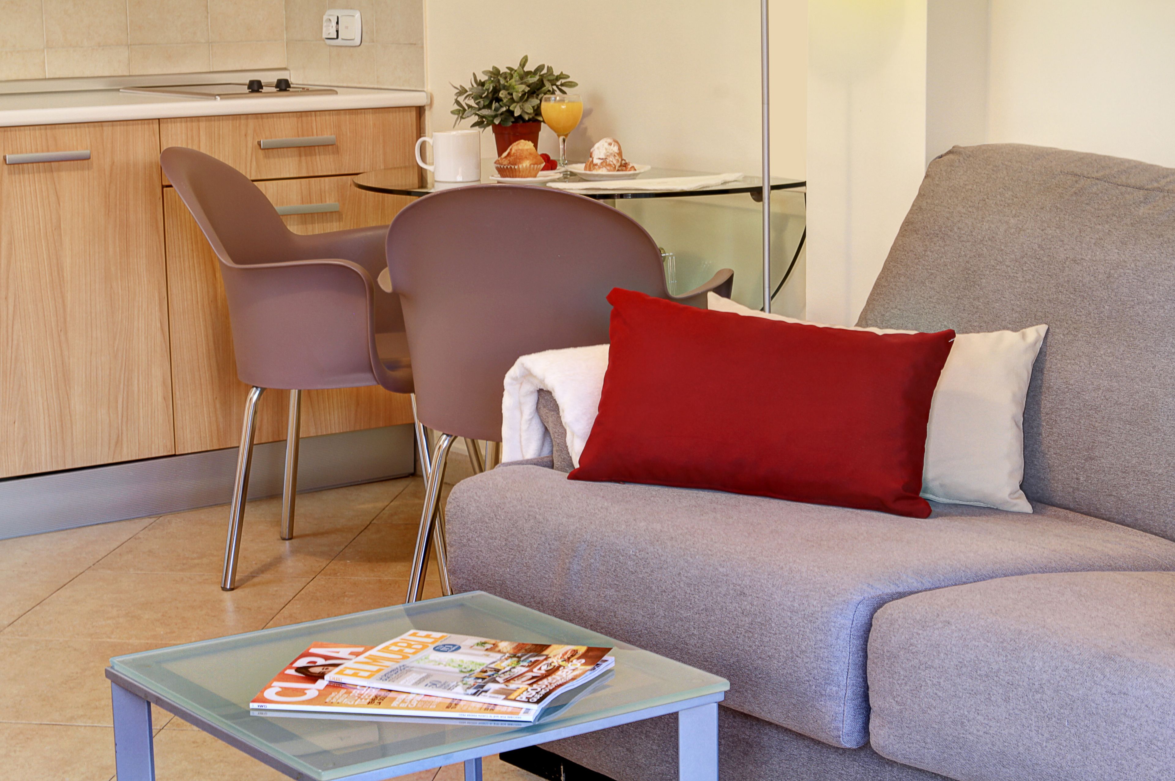 Uma sala de estar moderna com um sofá cinzento, um travesseiro vermelho, uma mesa de centro de vidro com revistas, e uma área de jantar com uma mesa branca, cadeiras e uma planta.