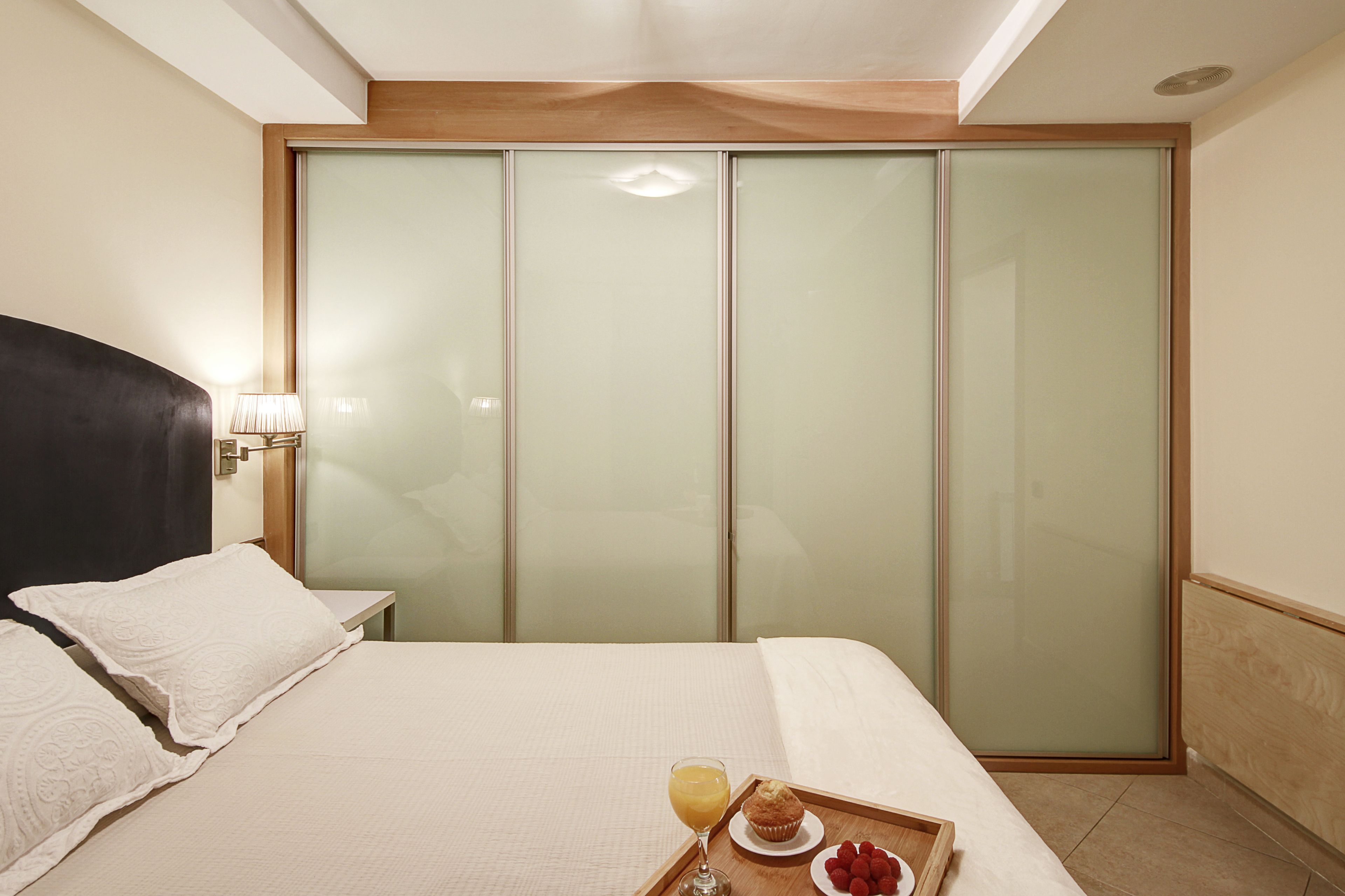 A hotel room with a bed, a nightstand, a glass partition, and a breakfast tray with a muffin and a glass of orange juice.