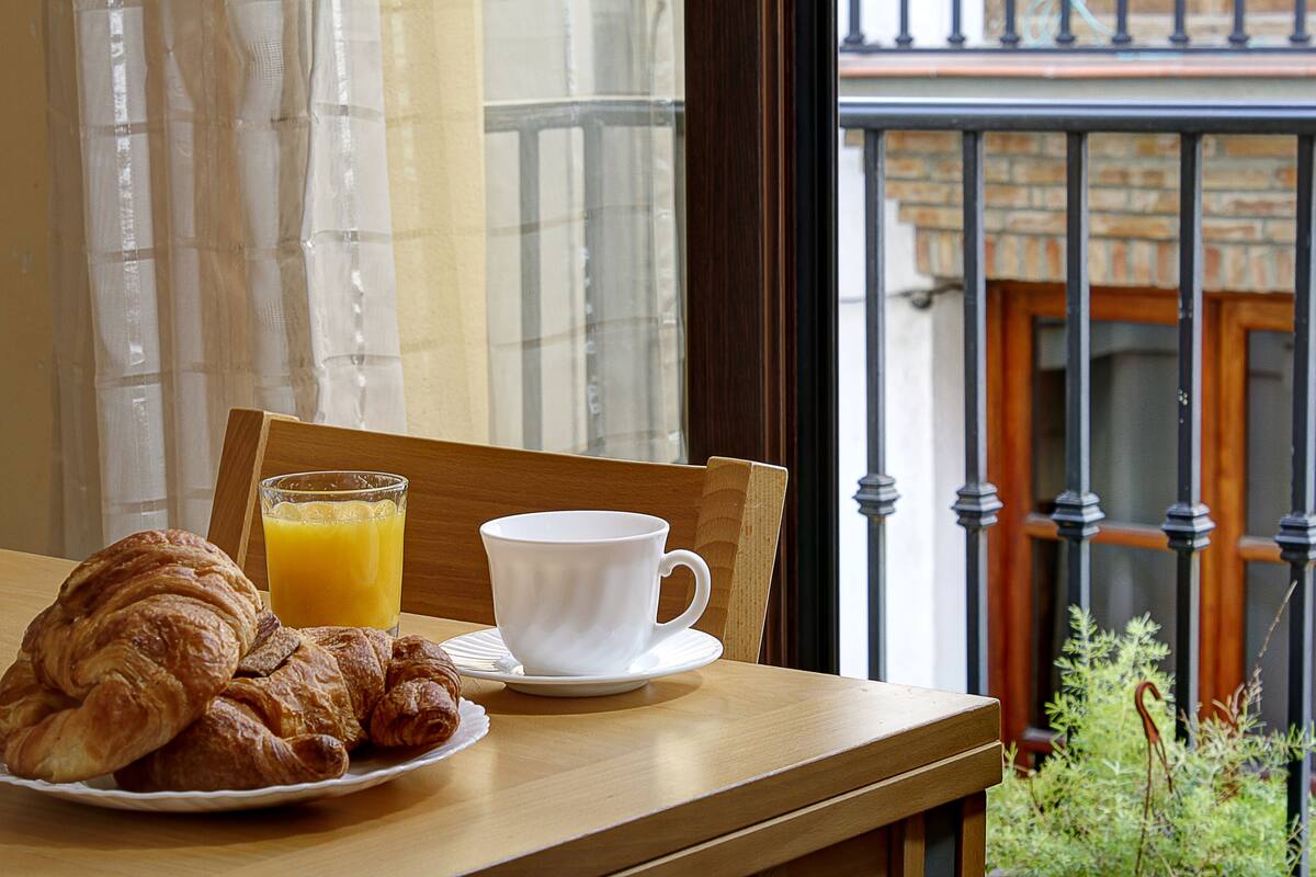 A breakfast setting with croissants and orange juice on a table, a white cup, and a view of a balcony with a railing and greenery.